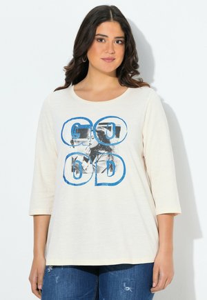 Femme aux cheveux bruns portant une chemise à manches longues crème avec le texte bleu "GOOD" et une impression photo en noir et blanc, associée à un jean bleu déchiré.
