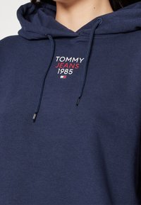 Felpa con cappuccio blu navy con cordini, caratterizzata dal logo "TOMMY JEANS 1985" ricamato in bianco e rosso. Texture morbida, design casual.