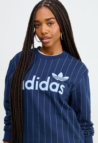 Marinblå sweatshirt med vita vertikala ränder och en stor ljusblå "adidas"-logotyp. Har rund halsringning och långa ärmar.