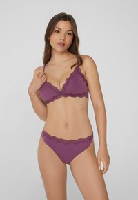Femme posant dans un soutien-gorge à volants violets et une culotte assortie, regardant la caméra sur un fond gris clair uni.
