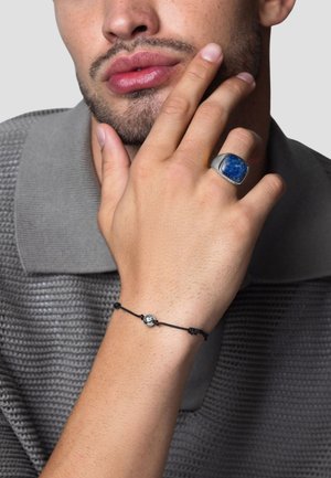 Silver ring with blue stone, worn on the right hand. Black cord bracelet features a small metallic ball. Grey textured knit shirt.