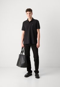 Zwarte polo met korte mouwen, groene logo-detail, gecombineerd met zwarte broeken en een zwarte leren totebag. Schoenen zijn zwart en stevig.