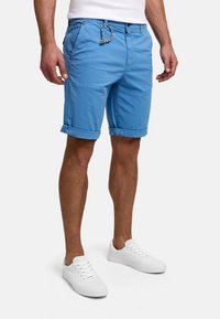 Man wearing cuffed blue shorts with drawstring and white sneakers, standing against a plain light background.