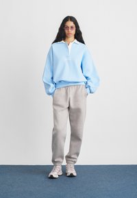 Žena v svetlom modrom pulóvri, šedých teplákach, ružových teniskách a ružovo zafarbených slnečných okuliaroch stojí na modrom koberci proti bielej stene.