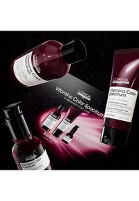 Trzy produkty do pielęgnacji włosów L'Oréal w burgundowych butelkach, z etykietami z czarnym tekstem. Towarzyszy im czarna karta promocyjna z szczegółami produktów.
