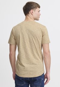 Tricou scurt bej cu maneci scurte, guler rotund și o croială lejeră, purtat de un model care se întoarce cu spatele, evidențiind designul de pe spate.