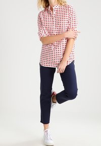 Nainen, jolla on päällään punavalkoinen ruudullinen paita, tummansiniset kapeat housut ja valkoiset lenkkarit, seisoo yhdellä jalalla yksinkertaista taustaa vasten.