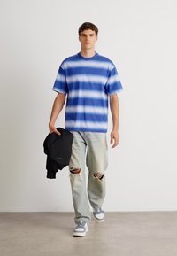 Camisa de rayas azul y blanca de manga corta, jeans claros desgastados, chaqueta negra, zapatillas grises, con desgastes visibles en los jeans.