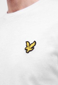 Aigle jaune brodé avec contour noir sur la poitrine d’un t-shirt blanc porté par une personne.