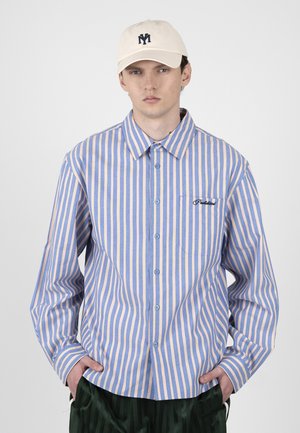 Fiatal férfi kék és bézs függőleges csíkos gombos ingben, bézs sapkában sötét logóval és sötétzöld nadrágban, kezek a zsebekben.