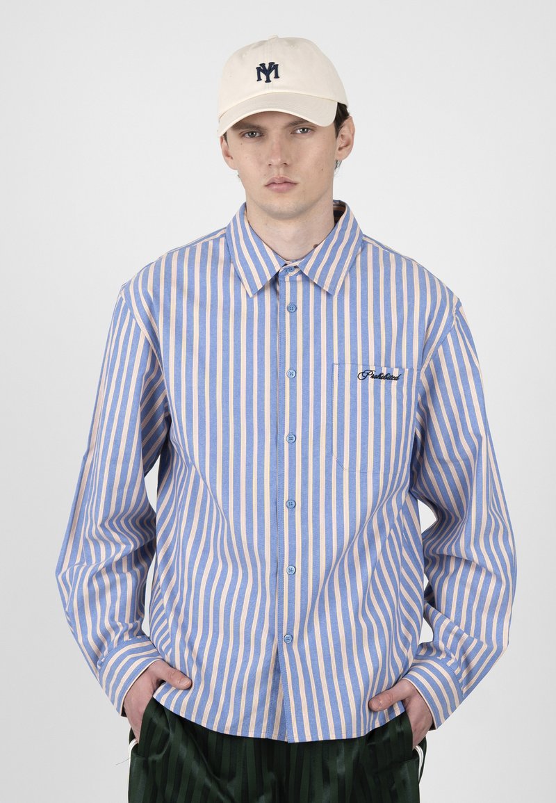 Jeune homme portant une chemise à boutons à rayures verticales bleu et beige, une casquette beige avec un logo foncé et un pantalon vert foncé, les mains dans les poches.