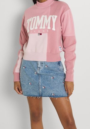 Femme portant un sweat-shirt rose Tommy Jeans et une jupe en jean bleue avec de petits logos brodés, debout contre un mur blanc.