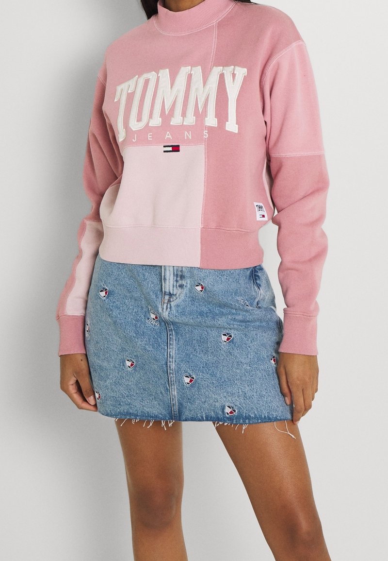 Femme portant un sweat-shirt rose Tommy Jeans et une jupe en jean bleue avec de petits logos brodés, debout contre un mur blanc.