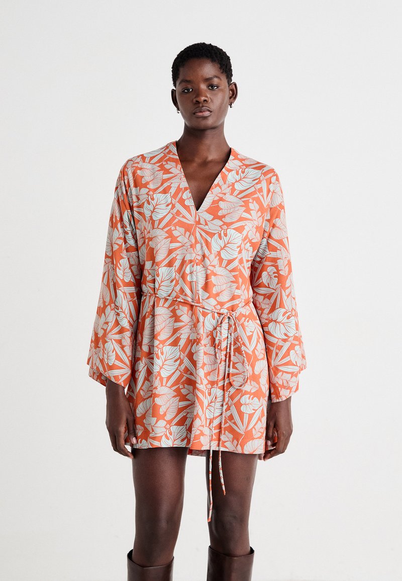 Robe florale orange avec un décolleté en V, des manches longues en forme de cloche et une taille à nouer. Présente des motifs de feuilles blanches et une texture de tissu douce.