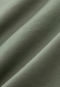 Grüner Stoff mit einer glatten Textur, diagonalen Falten und einem leichten Glanz. Das Material wirkt weich und eng gewebt.
