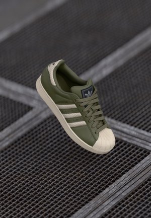 Green Adidas Superstar sneaker with white shell toe and stripes, floating above a textured metal grid surface.