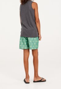 Persona che indossa una maglietta smanicata grigia scura, pantaloni corti verdi con motivi di palme gialle e infradito nere, in piedi di spalle.