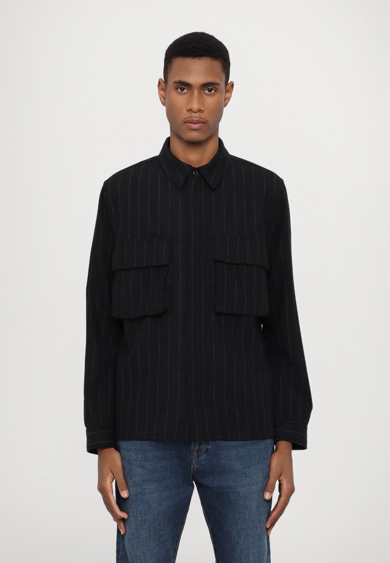 Svart randig jacka med två framfickor, knäppbar krage och lång ärm. Tyget verkar texturerat och passar bra med blå denimjeans.