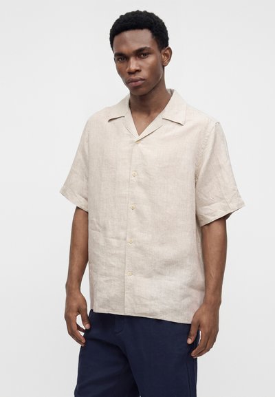 Homme portant une chemise beige à manches courtes boutonnée et un pantalon bleu foncé, debout devant un fond clair uni.