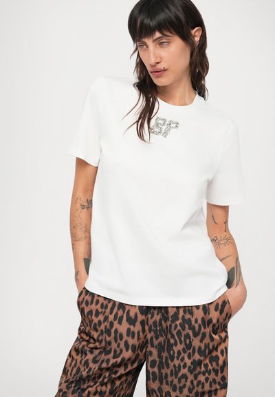 Valge lühikeste varrukatega t-särk, millel on sädelev "SP" logo. Koos pruunide leopardimustriga pükstega, millel on sile tekstuur.