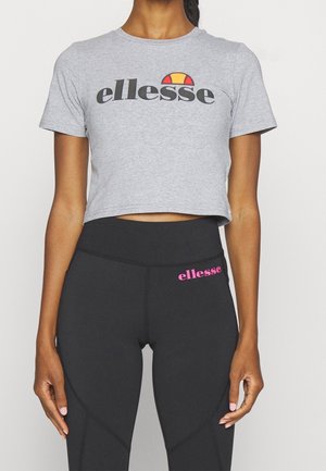 Kobieta nosząca szarą krótką koszulkę z logo „ellesse” i czarne legginsy z wysokim stanem z różowym logo „ellesse” na lewym biodrze.