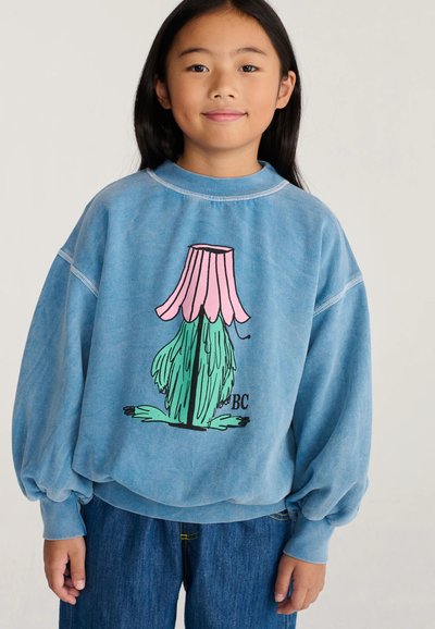 Fille aux longs cheveux noirs portant un sweat-shirt bleu avec un motif abstrait de fleur de dessin animé rose et verte et un jean bleu.