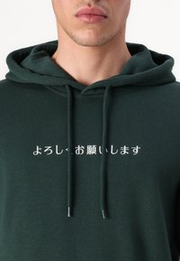 Grüne Kapuzenpullover aus weichem Material, mit weißem japanischen Text auf der Vorderseite. Mit Kordeln und einem lockeren Schnitt.