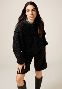 Giacca bomber nera con motivo floreale testurizzato, paillettes lucide e polsini a costine, abbinata a pantaloncini neri su misura e stivali al ginocchio.