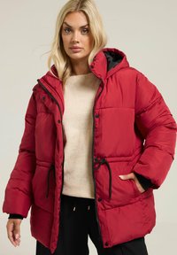 Veste matelassée rouge avec capuche, dotée d'une doublure noire, d'une fermeture à pression à l'avant et d'une taille ajustable par cordon. Le tissu semble lisse avec une texture matelassée.
