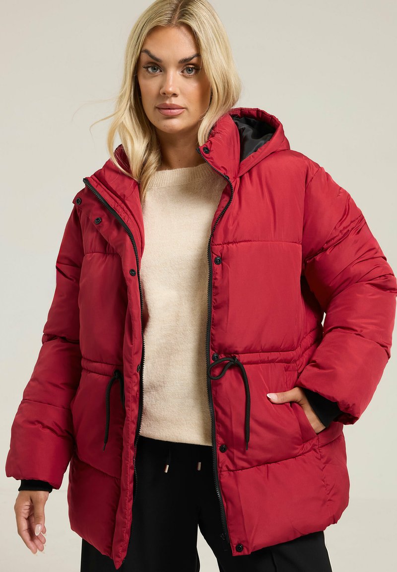 Veste matelassée rouge avec capuche, dotée d'une doublure noire, d'une fermeture à pression à l'avant et d'une taille ajustable par cordon. Le tissu semble lisse avec une texture matelassée.