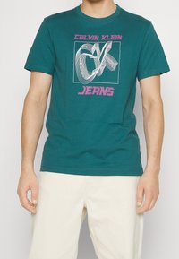 Turkusowa bawełniana koszulka z grafiką zawierającą napis "CALVIN KLEIN" oraz "JEANS" w różowym kolorze, z krótkim rękawem i klasycznym okrągłym dekoltem.