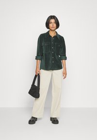Groene corduroy shirt met twee borstzakken, knoopsluiting, opgerolde mouwen; gecombineerd met crèmekleurige wijde broek en een zwarte tas. Zwarte chunky laarzen.