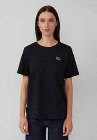 Zwarte katoenen t-shirt met korte mouwen, featuring een klein bronzen pretzel-vormig embleem op het linkerbovenbeen, met een relaxed fit.