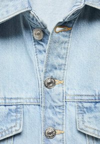 Veste en denim bleu clair avec une fermeture à boutons-pressions argentés, un col pointu et deux poches poitrine avec couture apparente.