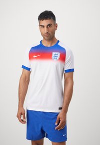 Hvid fodboldtrøje med rød og blå gradientdesign, Nike-logo og Englands våbenskjold. Sammen med blå shorts. Glat, åndbart stof.
