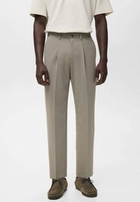 Mandlig model iført beige plisserede bukser, brune ruskind snørebåndssko og en off-white T-shirt med korte ærmer foran en ensfarvet baggrund.