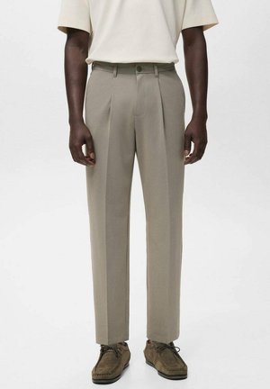 Male model wearing beige pleated trousers, brown suede lace-up shoes, and off-white short-sleeve shirt against plain background.