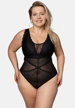 Smiling woman with long blonde hair wearing black lace and mesh bodysuit, standing against a plain light background.