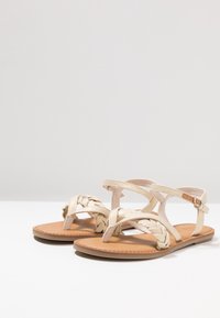 Vaaleat beiget litteät sandaalit, joissa on punottu varvasremmi ja useita ristiin meneviä remmejä. Luonnon nahkainen pohja ja vaaleanruskea kumipohja.