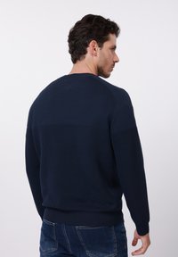 Pull en tricot bleu marine avec texture côtelée, col rond et poignets ajustés. Présente un motif à rayures horizontales sur le haut du dos.