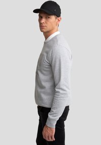 Mand iført grå langærmet skjorte, sort kasket og sorte bukser står sideværts mod en ensfarvet grå baggrund.