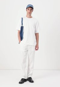 T-shirt bianca oversize, pantaloni bianchi in lino, mocassini neri, borsa a tracolla in denim blu, cappellino blu, in piedi contro uno sfondo chiaro uniforme.