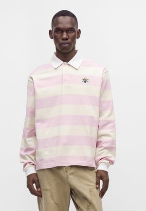 Giovane uomo che indossa una polo a maniche lunghe a righe rosa e bianche con un piccolo ricamo di teschio e cono gelato.
