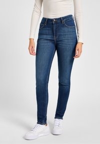 Vaqueros pitillo de mezclilla azul oscuro con cintura alta, que cuentan con bolsillos delanteros, cierre de botón y detalles de costura. Combinados con zapatillas blancas.