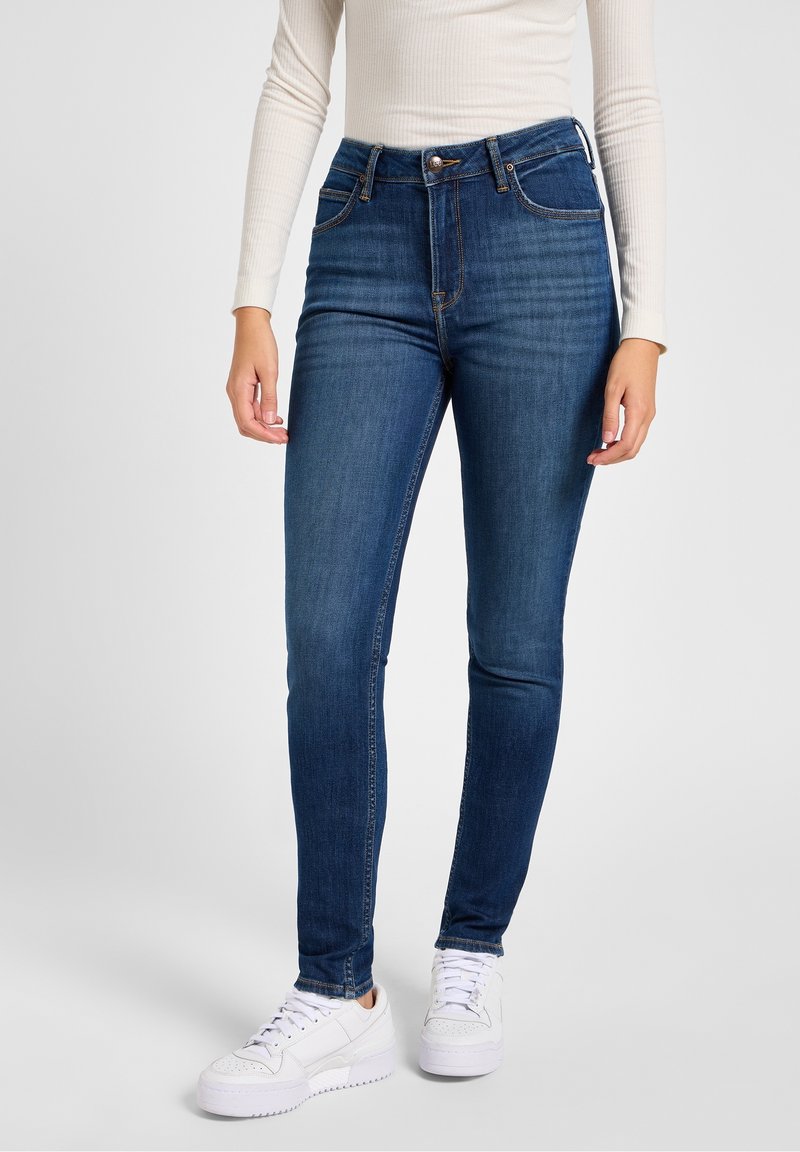 Vaqueros pitillo de mezclilla azul oscuro con cintura alta, que cuentan con bolsillos delanteros, cierre de botón y detalles de costura. Combinados con zapatillas blancas.