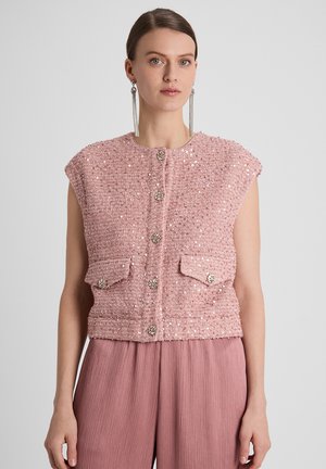 Vrouw draagt een mouwloze roze cardigan met paillettenstructuur en decoratieve knopen en bijpassende geplooide wijde broekspijpen, staand tegen een effen achtergrond.