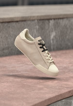 Bézs színű velúr alacsony szárú sportcipő krémszínű orrkarral, fekete és krémszínű fűzők keverékével, rózsaszín kőlépcsők felett lebegve.