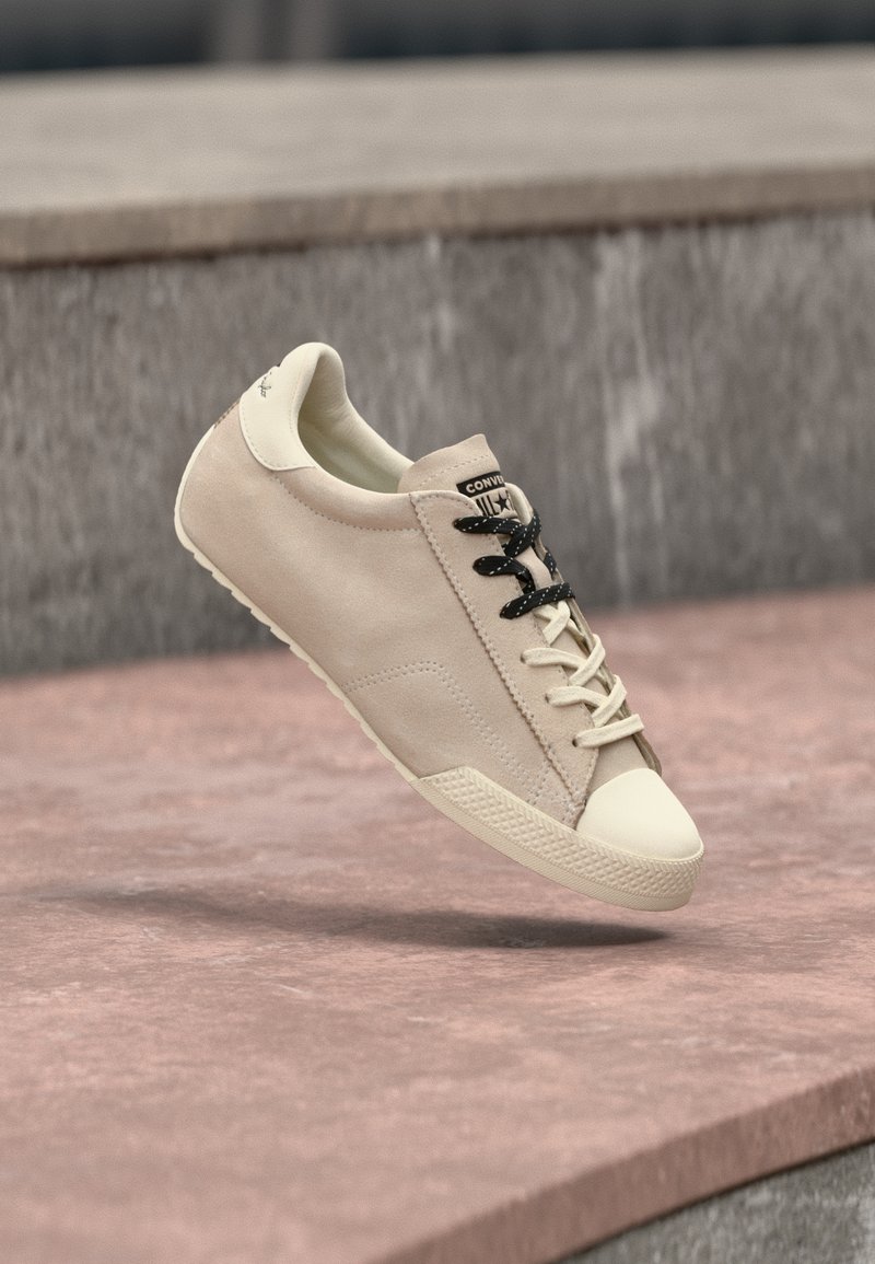 Μπεζ σουέτ αθλητικό παπούτσι χαμηλής κορυφής με εκρού προστατευτικό στη μύτη, κορδόνια σε μίξη μαύρου και εκρού, αιωρούμενο πάνω από ροζ πέτρινα σκαλοπάτια.