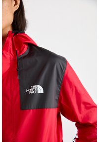 Czerwona kurtka przeciwdeszczowa z czarnym panelem na ramieniu, z logo z przodu w kolorze białym. Teksturowany materiał z regulowanym kapturem i sznurkami.