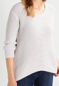 Pull en maille gris clair avec col en V, texture côtelée et manches longues, présentant un ourlet asymétrique et des coutures verticales marquées.